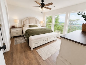 A bedroom with a bed, bedside tables, a nightstand, a bench, a ceiling fan, and a view through large windows. - Bluff View Overlooking Sequatchie Valley. 2 King Beds + Hot Tub (Graysville)