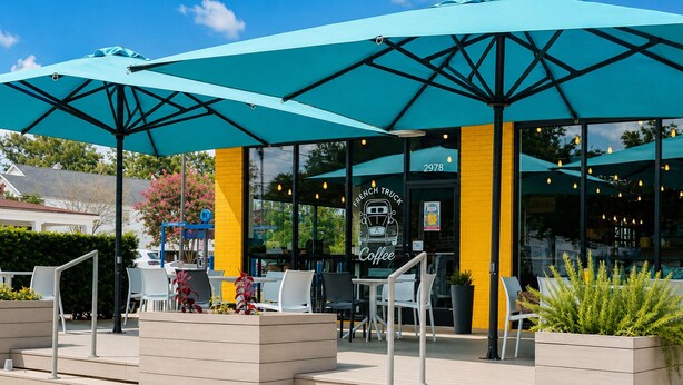 Een terras met tafels en stoelen onder grote parasols, een winkel met een bordje 'French Truck Coffee' en een geel bakstenen muur.