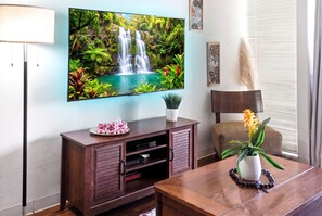 A living room with a flat-screen TV mounted on the wall, a wooden entertainment unit, a dining table with a vase of flowers, and a window with blinds. - Kihei Holiday 215 (Kihei)