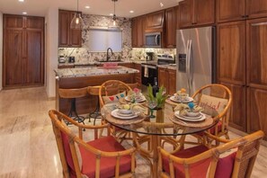 A kitchen with wooden cabinets, a marble countertop, a stainless steel refrigerator, and a dining area with wicker chairs and a glass table. - Beachfront Beautiful 2 Bedroom 2 Bathroom Condo near surf break (San  Jose del Cabo)