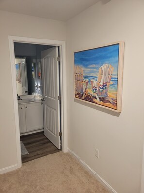 A hallway with a door leading to a bathroom, a painting of beach chairs on the wall, and a carpeted floor. - Coastal Retreat w/Resort Pools & Shuttle to Beach (Millville)
