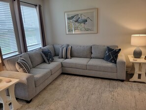 Ein Wohnzimmer mit einem grauen Ecksofa, einem gerahmten Vogelgemälde, einer Lampe und einem kleinen Tisch.