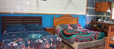 Una habitación con dos camas, cabecero de madera, piso de baldosas y una zona de cocina con armarios y un lavamanos.