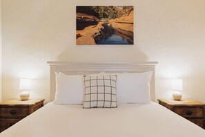 Ein Bett mit weißen Leinentüchern, einem karierten Kopfkissen und zwei Nachttischlampen auf Holznachttischen.