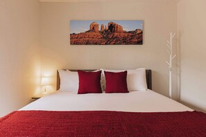 Une chambre d’hôtel avec un lit, une table de chevet avec une lampe, un plaid rouge et un tableau représentant un paysage désertique accroché au mur.