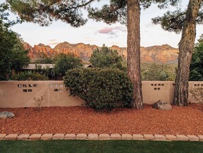 A landscaped area with a rock wall, a small bush, and a view of red rock formations in the background. - Charming Home w/ Casita, Hot Tub, Red Rock Views (Sedona)