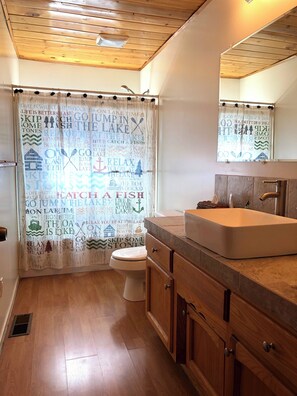 A bathroom with a wooden floor, a white toilet, a wooden vanity with a sink, and a shower curtain with text and graphics. - 3-bedroom cabin in gorgeous South Lake Tahoe with AC (South Lake Tahoe)