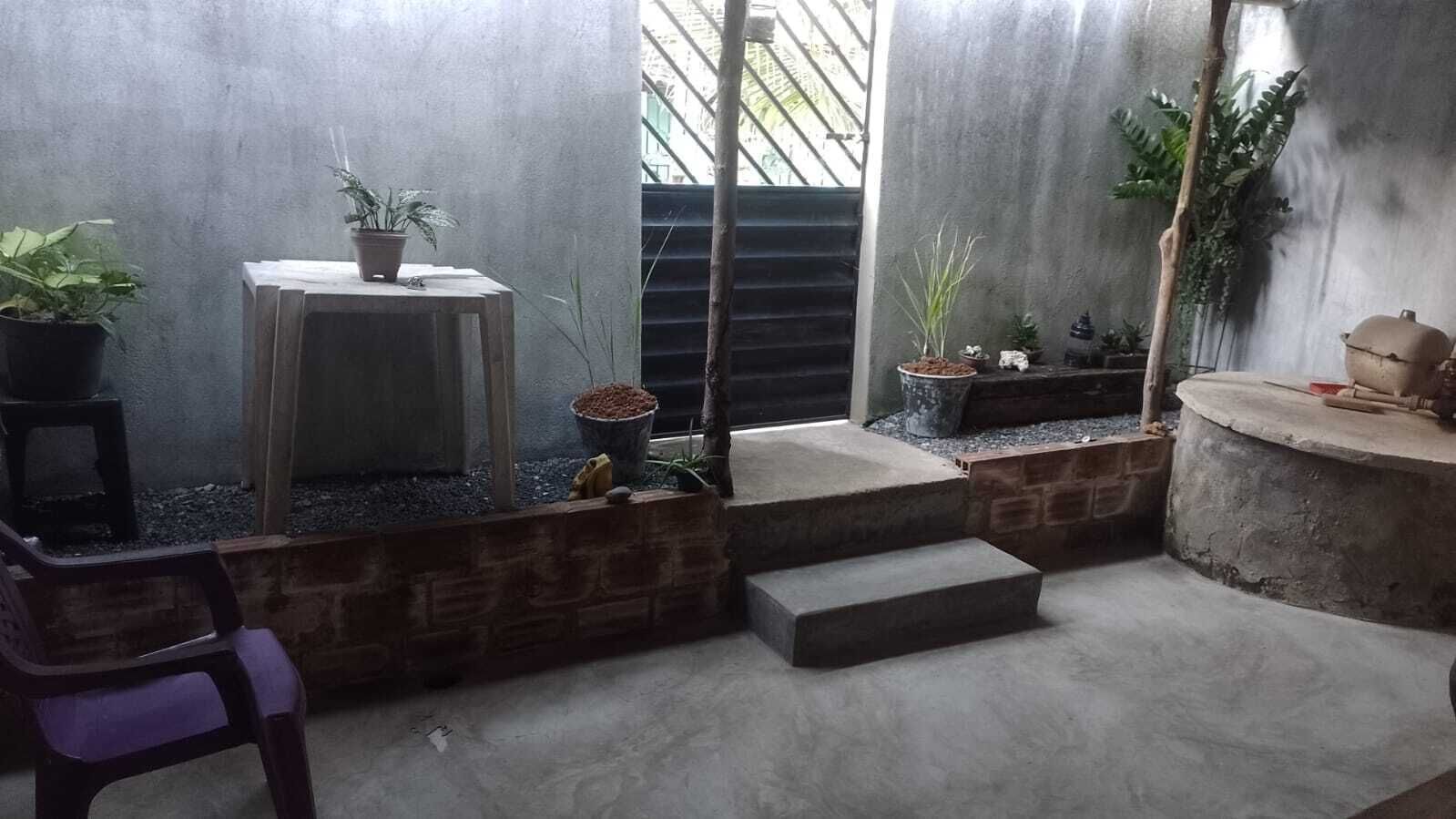 A small, enclosed outdoor area with a concrete floor, a wooden stool, a purple chair, and several potted plants.