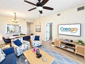 A modern living room with a ceiling fan, a dining area with a table and chairs, a television displaying 'Casago Vacation Rentals,' and a bookshelf with various items. - Coastal Luxury at Surf Club | Direct Oceanfront | Resort-Style Amenities (Palm Coast)