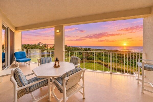 Un balcon avec une table et des chaises, une vue sur le coucher de soleil sur l’océan et une balustrade.
