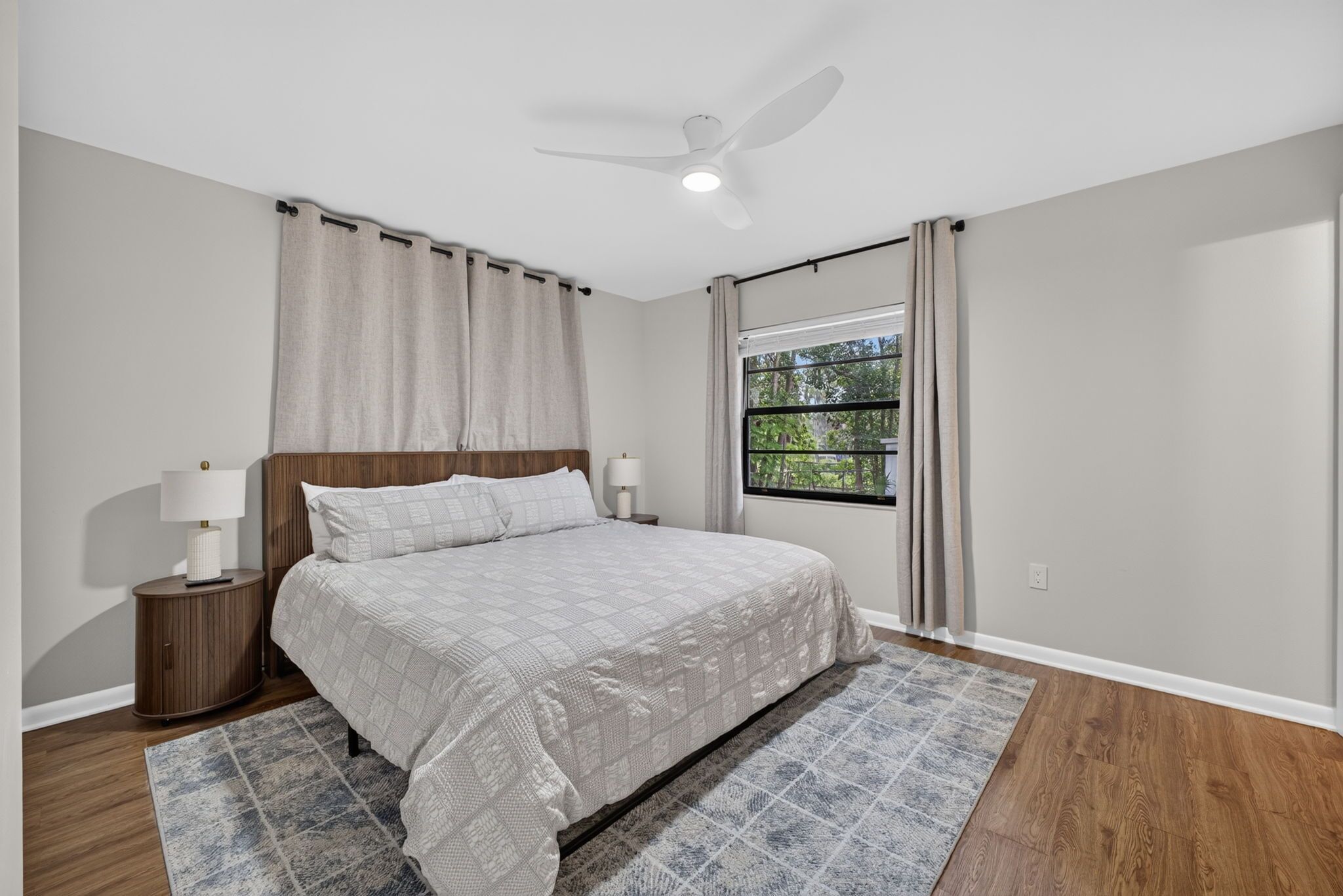 Une chambre à coucher comprenant un lit, des tables de chevet, un ventilateur de plafond, une fenêtre avec des rideaux et une vue sur la verdure à l’extérieur.