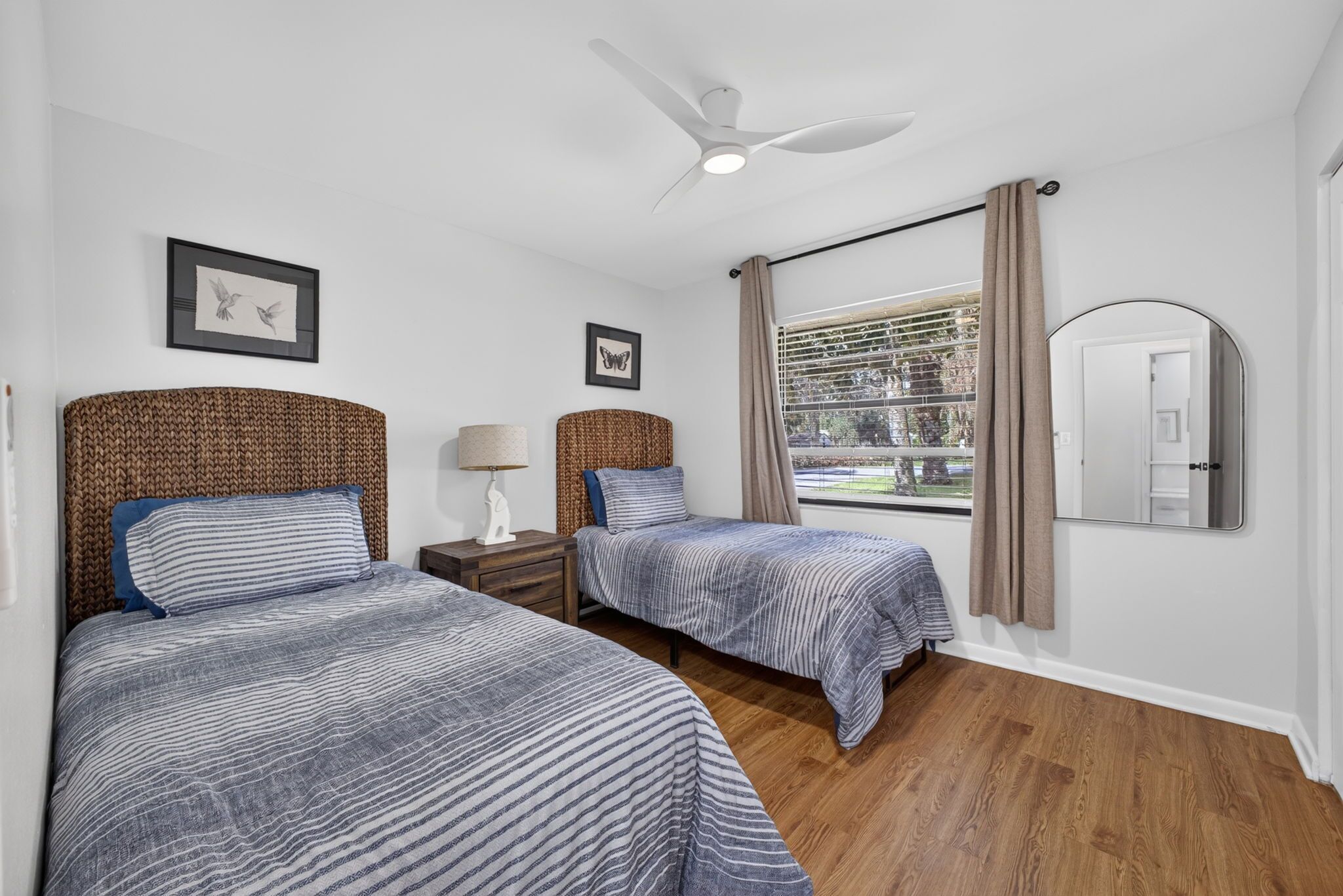 A hotel room with two beds, a ceiling fan, a window with curtains, and a mirror.