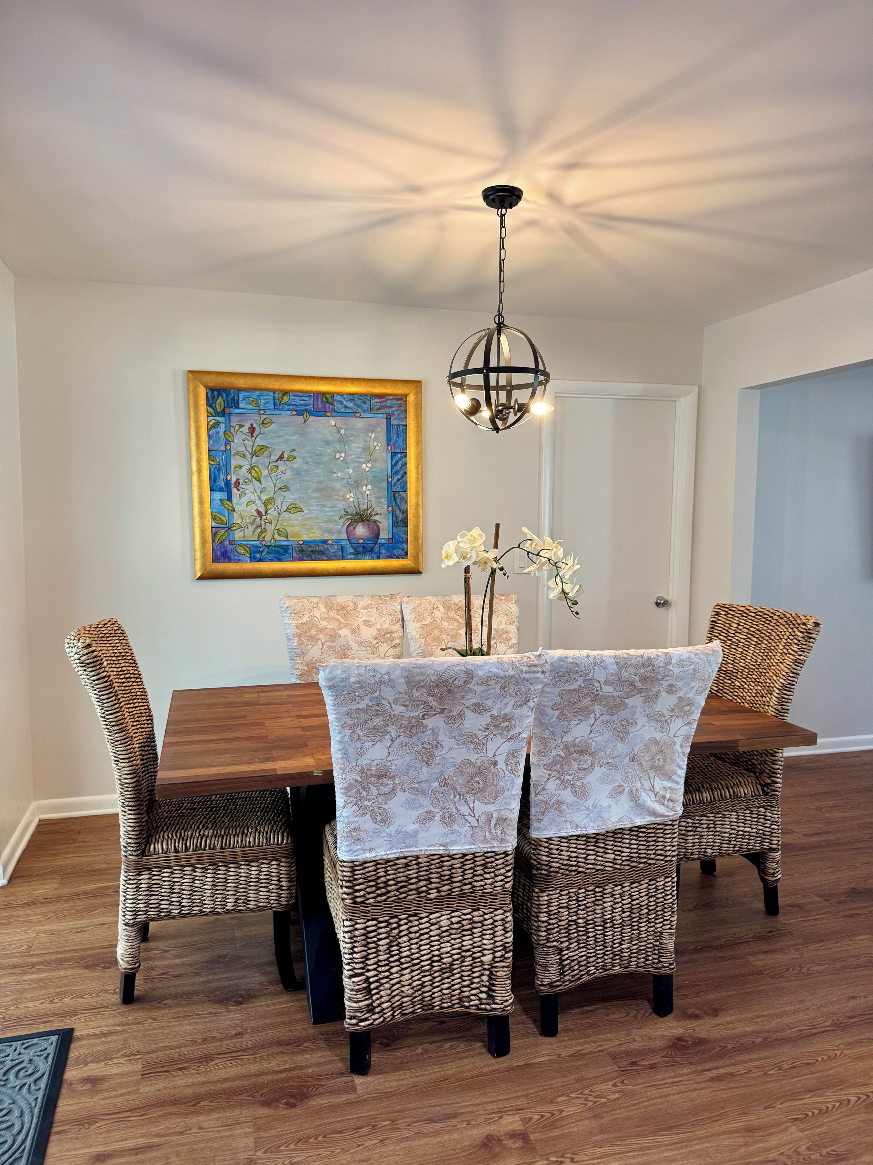 A dining area with a wooden table, wicker chairs, a floral painting, and a hanging light fixture.
