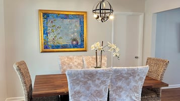 A dining area with a wooden table, wicker chairs, a floral painting, and a hanging light fixture.