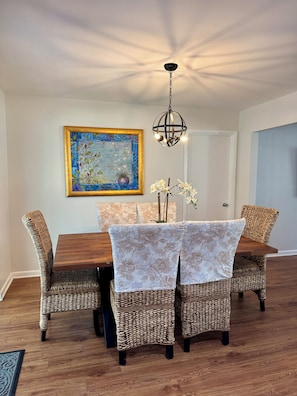 A dining area with a wooden table, wicker chairs, a floral painting, and a hanging light fixture.
