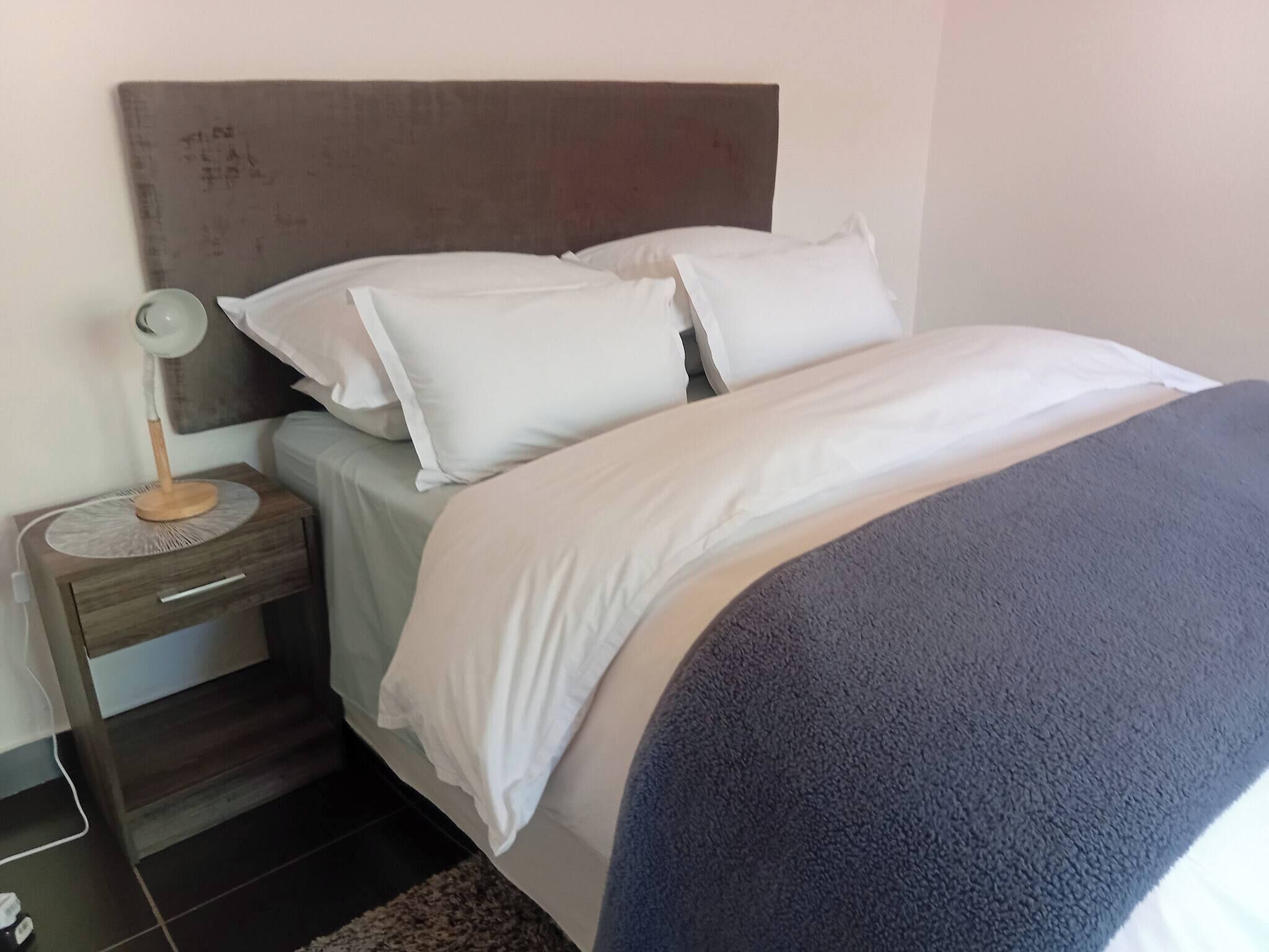Una cama bien hecha con ropa blanca y una manta azul, una mesita de noche con una lámpara y un cabecero.