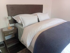 A neatly made bed with white bedding and a blue blanket, a bedside table with a lamp, and a headboard. - Executive Garden Flat in Plk (Polokwane)