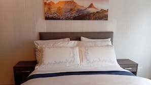 Un lit avec une literie blanche et un élément décoratif bleu, une table de chevet et un tableau représentant un paysage de montagne accroché au mur.