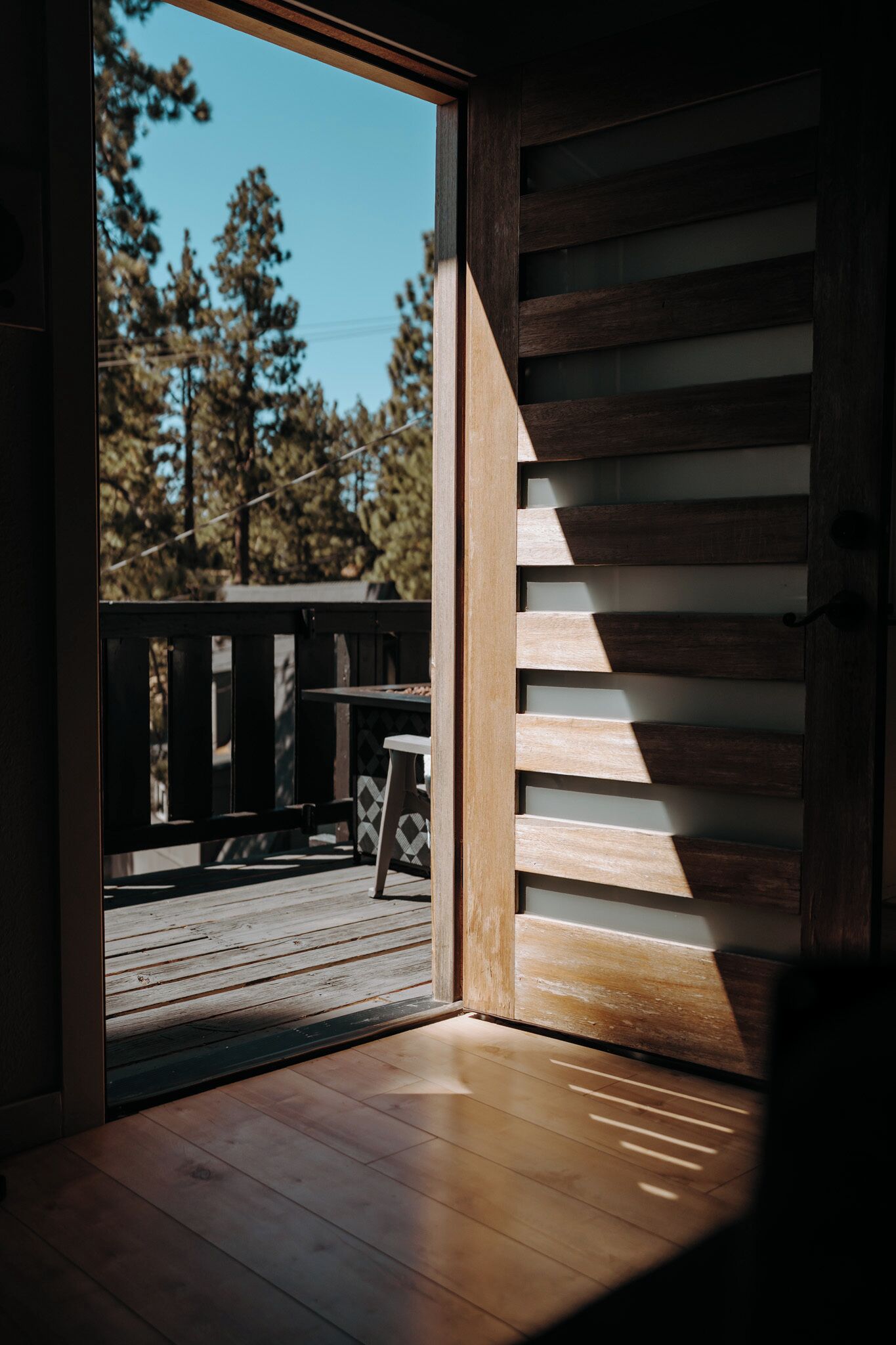 A room with a wooden floor, a view of a deck with a table and chairs, and a large window with a view of trees.
