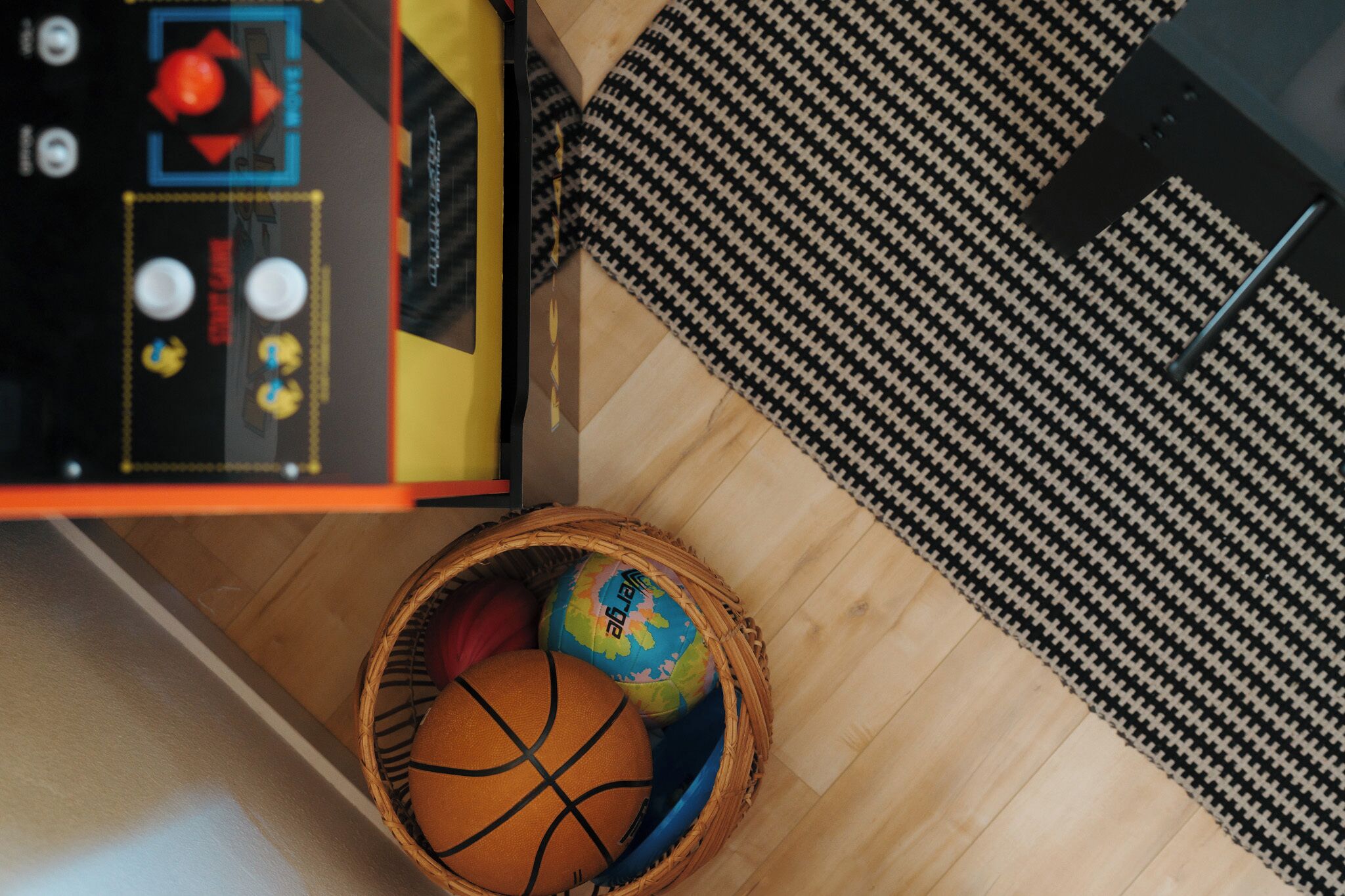 A room with a herringbone-patterned rug, a wooden floor, and a basket containing a basketball and a ball with a colorful design.