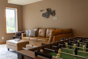 A living room with a foosball table, a brown leather sofa, and a window.