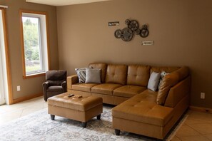 A living room with a sectional sofa, a single armchair, and a coffee table. There are wall decorations and a window with a view. - Spacious Madawaska River Cottage with Hot Tub, Games Room & Dock (Brudenell, Lyndoch and Raglan)
