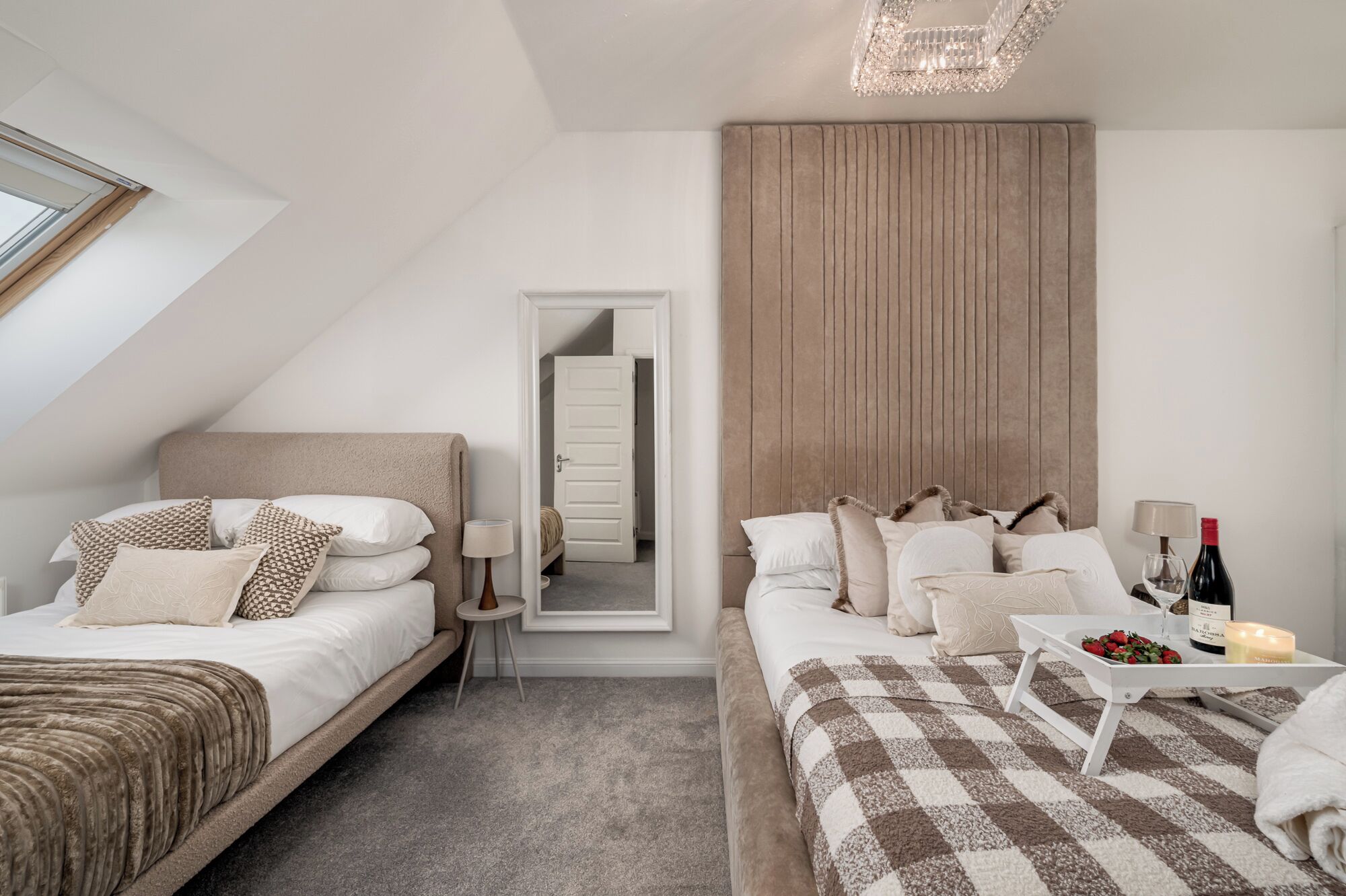 Une chambre d’hôtel moderne avec deux lits, une grande fenêtre, un luminaire au plafond et une table de chevet sur laquelle se trouvent une bouteille et une bougie.
