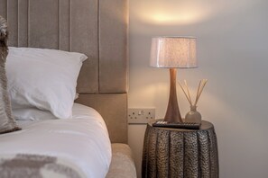 Ein ordentlich bezogenes Bett mit Kissen, eine Nachttischlampe mit einer strukturierten Schirm und ein kleiner Tisch mit Fernbedienung und Reed-Diffusor.