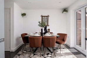 A dining area with a table set for four, surrounded by brown chairs, a black vase with plants, and a chalkboard sign on the wall. - Chic 4BR Home | Garden | Free Parking | Maidstone (Kent)