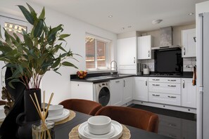 A modern kitchen with white cabinets, a built-in oven, and a dining area with a table set for two. - Chic 4BR Home | Garden | Free Parking | Maidstone (Kent)