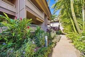 A hotel with a covered outdoor area, a concrete pathway, and various plants and flowers. - Kihei Bay Vista B102 (Kihei)
