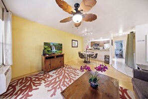 A living room with a ceiling fan, a television, a dining table with chairs, a kitchen in the background, and a sofa. - Kihei Bay Vista B102 (Kihei)