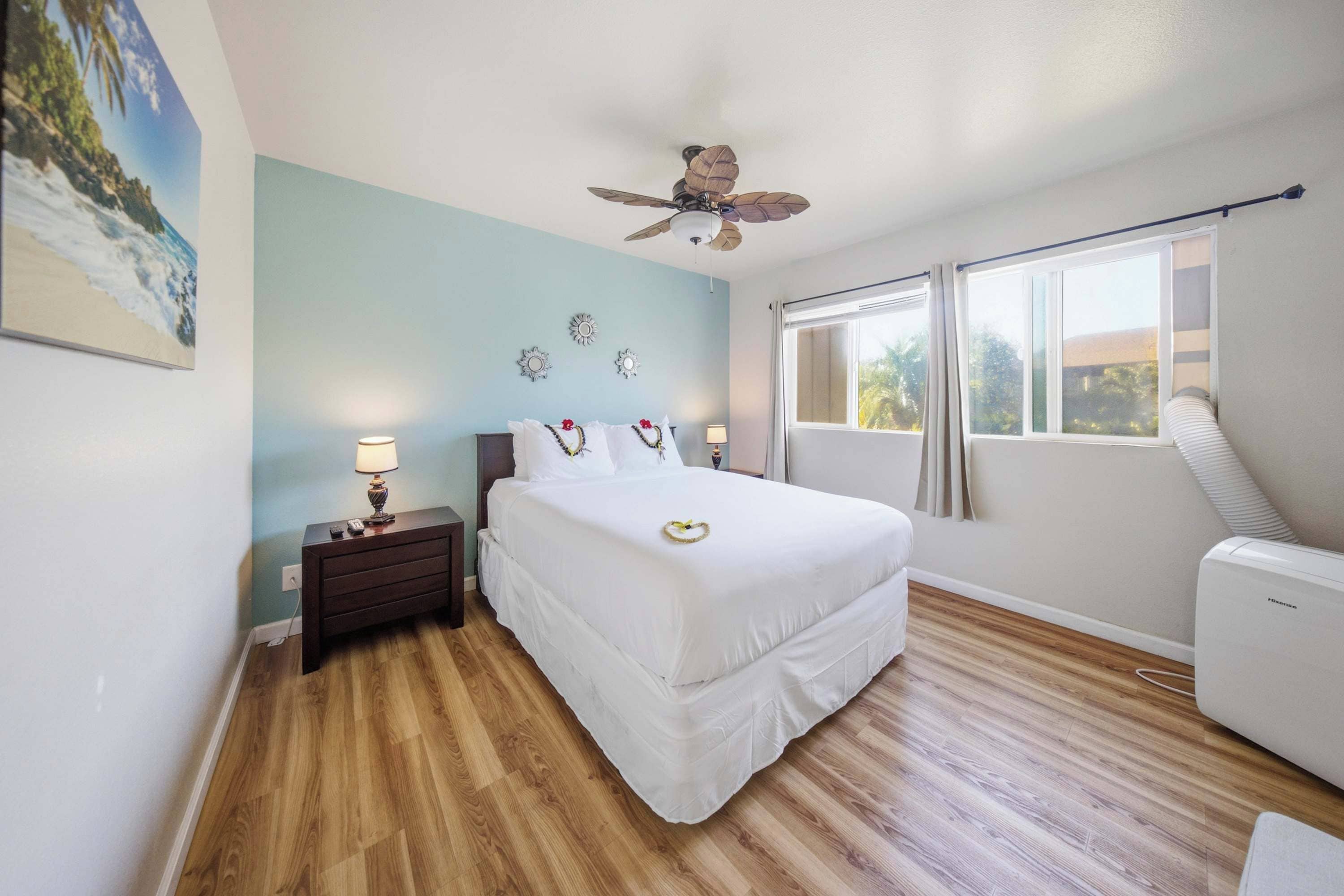 A bedroom with a bed, bedside tables, a ceiling fan, a window with curtains, and a wall-mounted air conditioning unit.