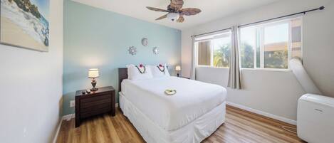 A bedroom with a bed, bedside tables, a ceiling fan, a window with curtains, and a wall-mounted air conditioning unit.