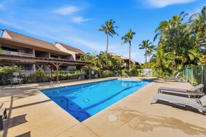 A swimming pool with lounge chairs and a covered area, surrounded by palm trees and a building with a sloped roof. - Kihei Bay Vista B102 (Kihei)