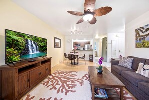 A living room with a flat-screen TV, wooden furniture, a ceiling fan, and a kitchen in the background. - Kihei Bay Vista B102 (Kihei)