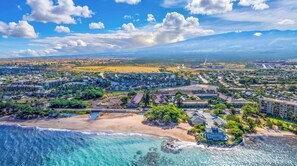 A coastal residential area with beachfront properties, a large building complex, and a mountain range in the background. - Kihei Bay Vista B102 (Kihei)