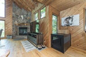 A cabin interior with a stone fireplace, wooden walls, a large window, and a mounted artwork.
