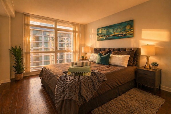 Une chambre à coucher avec un grand lit, une table de chevet, une lampe, un tableau et une fenêtre avec des rideaux.