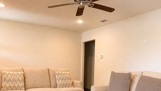 Una sala de estar con una mesa de centro marrón oscuro, dos sofás color beige con cojines estampados y un ventilador de techo.