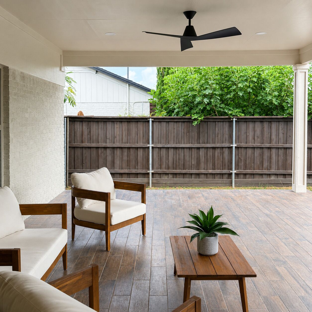 Un patio techado con ventilador de techo, una mesa de madera con planta en maceta y una zona de estar con sofá y sillones acolchados.