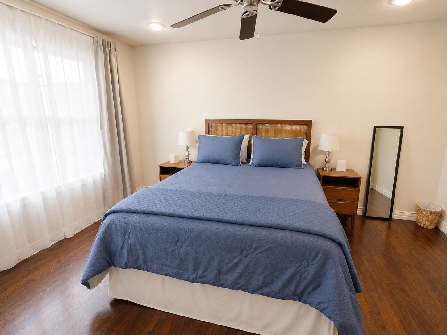 A bedroom with a bed, two bedside tables with lamps, a ceiling fan, a mirror, and a window with curtains.