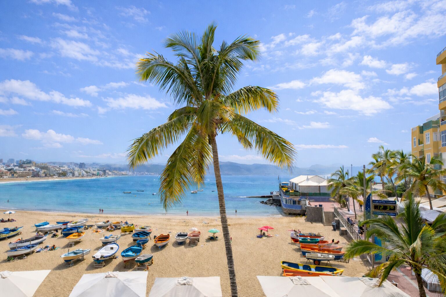 Ein Strand mit einer Palme, Booten, Sonnenschirmen und einem Gebäude mit einer digitalen Uhr.