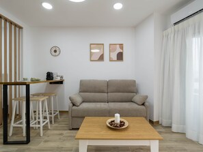Ein Wohnzimmer mit Sofa, Couchtisch, Esstisch mit Stühlen, einer Uhr und zwei gerahmten Bildern an der Wand.