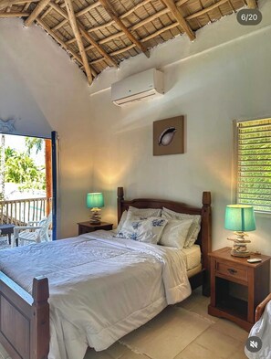 Quarto com cama de madeira, duas mesas de cabeceira com luminárias, ventoinha no teto, ar condicionado fixado na parede e janela com vista para o verde.