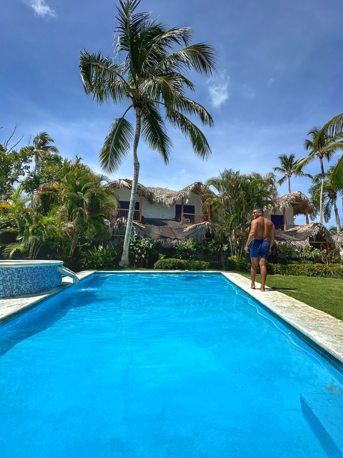 Une piscine avec une personne debout au bord, entourée de palmiers et d’un bâtiment au toit de chaume.
