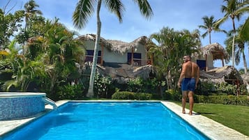 Une piscine avec une personne debout au bord, entourée de palmiers et d’un bâtiment au toit de chaume.