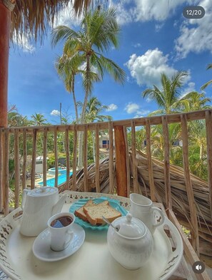 Varanda com mesa posta para o café da manhã, com vista para uma piscina e palmeiras.