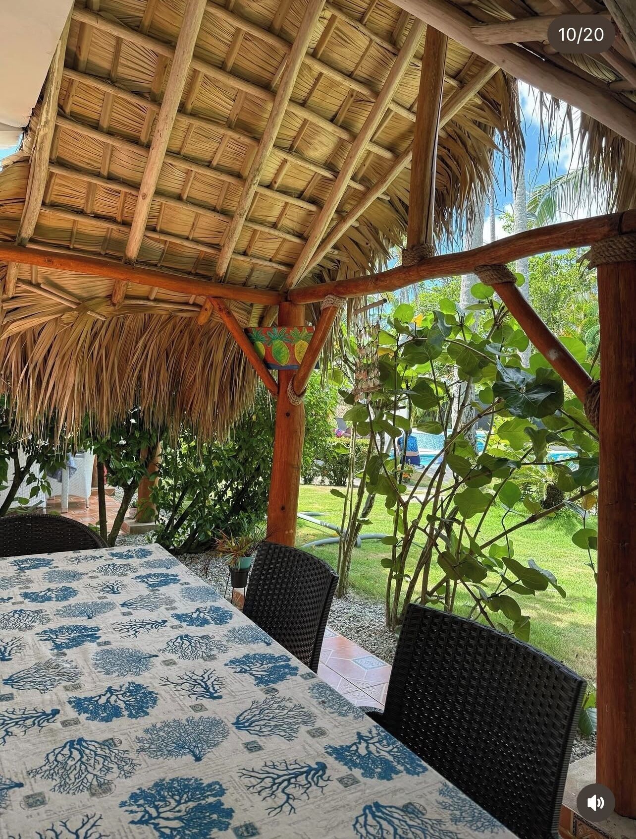 Un espace extérieur abrité, doté d’un toit de chaume, de chaises en osier et d’une table recouverte d’une nappe à motifs bleus et blancs.