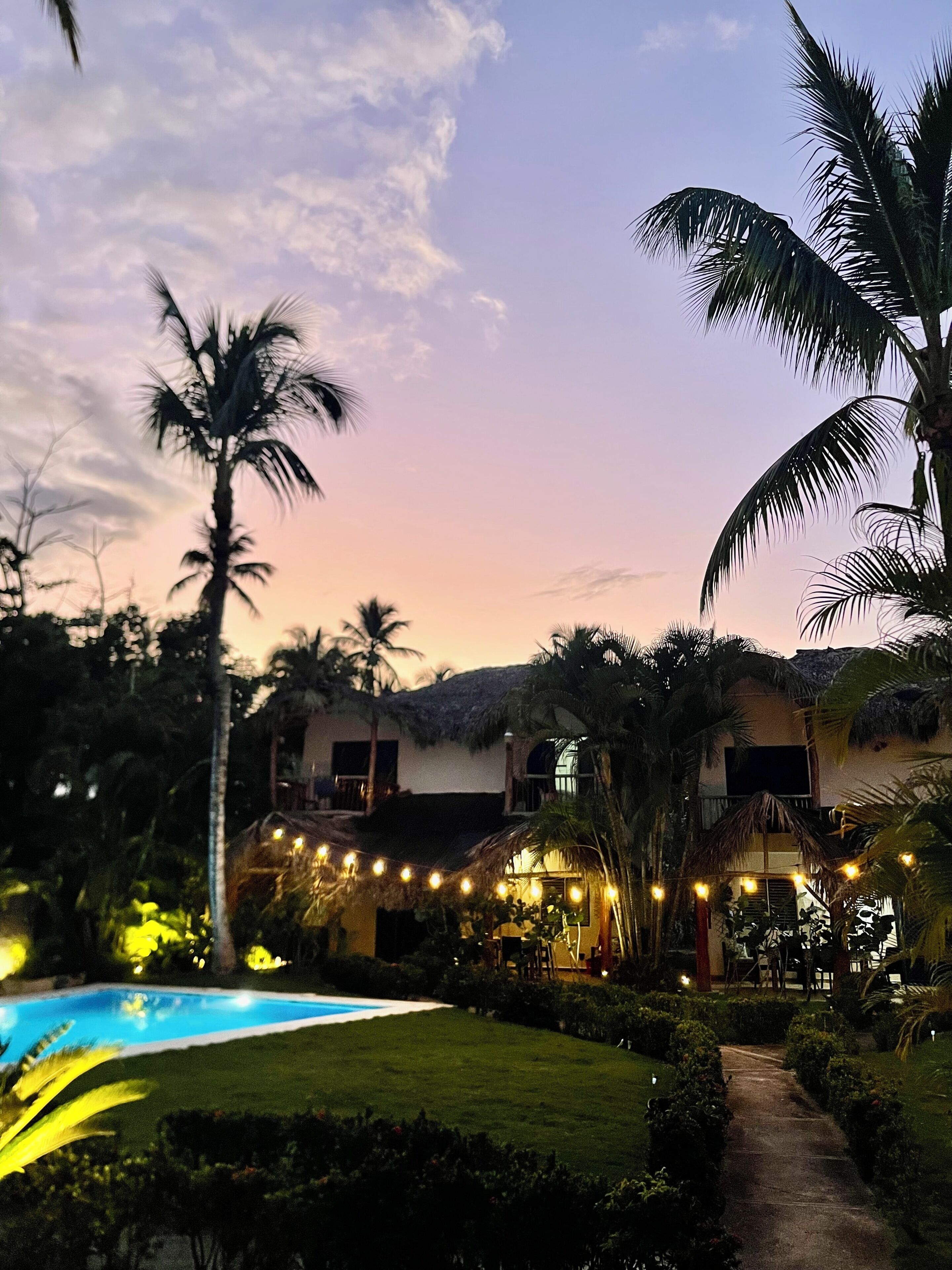 A pool area with a pathway leading to a building with a thatched roof, surrounded by palm trees and greenery.
